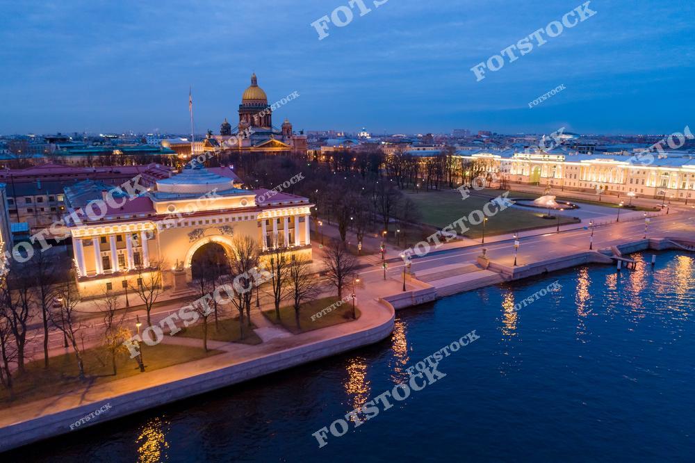 На фотографии запечатлено здание Адмиралтейства, освещенное вечерними огнями, с его характерным шпилем. На заднем плане возвышается Исаакиевский собор, величественно выделяющийся в городском пейзаже. Справа видна часть Дворцовой набережной, отражающей свет фонарей в воде Невы. Вид с высоты раскрывает красоту и гармонию архитектурных памятников Санкт-Петербурга.